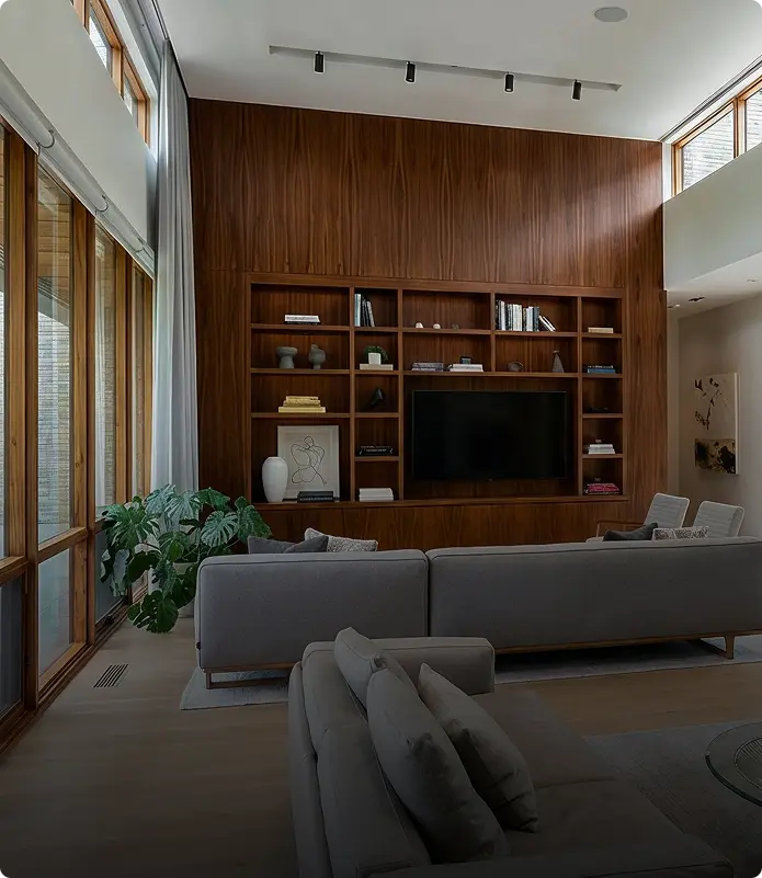 A modern living room with tall windows, wooden wall paneling, built-in shelves around a TV, gray sofas, a potted plant, and light wood flooring. Sunlight streams in, creating a bright and inviting space.